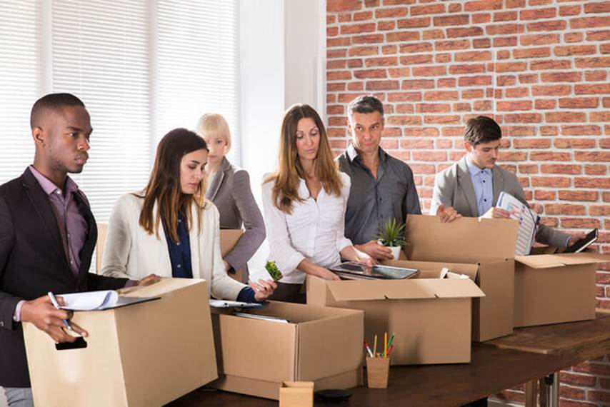 Employees pack belongings into cardboard boxes for a layoff. Several people stand at a table with open boxes, packing items from their desks.